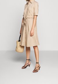 Beige skjortklänning med korta ärmar, knutet midjedetalj, matchad med svarta sandaler med remmar, håller en vävd rund väska.