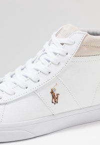 Valkoiset korkeavartiset tennarit, joissa on kanvaspinta, beige mokkanahka-akseli, litteät nauhat ja brodeerattu logo, jossa on polo-pelaaja hevosella.