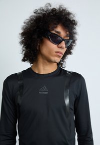 Čierna košeľa s dlhým rukávom a lesklými čiernymi akcentmi s logom Adidas. Model nosí elegantné, ostré čierne slnečné okuliare a má kučeravé vlasy.