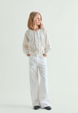 Joven de pie con las manos en los bolsillos, vistiendo una blusa blanca suelta, pantalones anchos blancos y zapatos negros con tachuelas, mirando de lado.