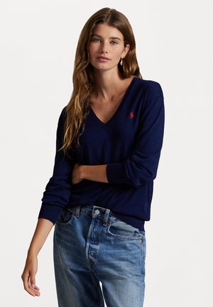 Young woman with long wavy hair wearing a navy blue V-neck sweater with a red logo and light blue denim jeans, standing against a white background.