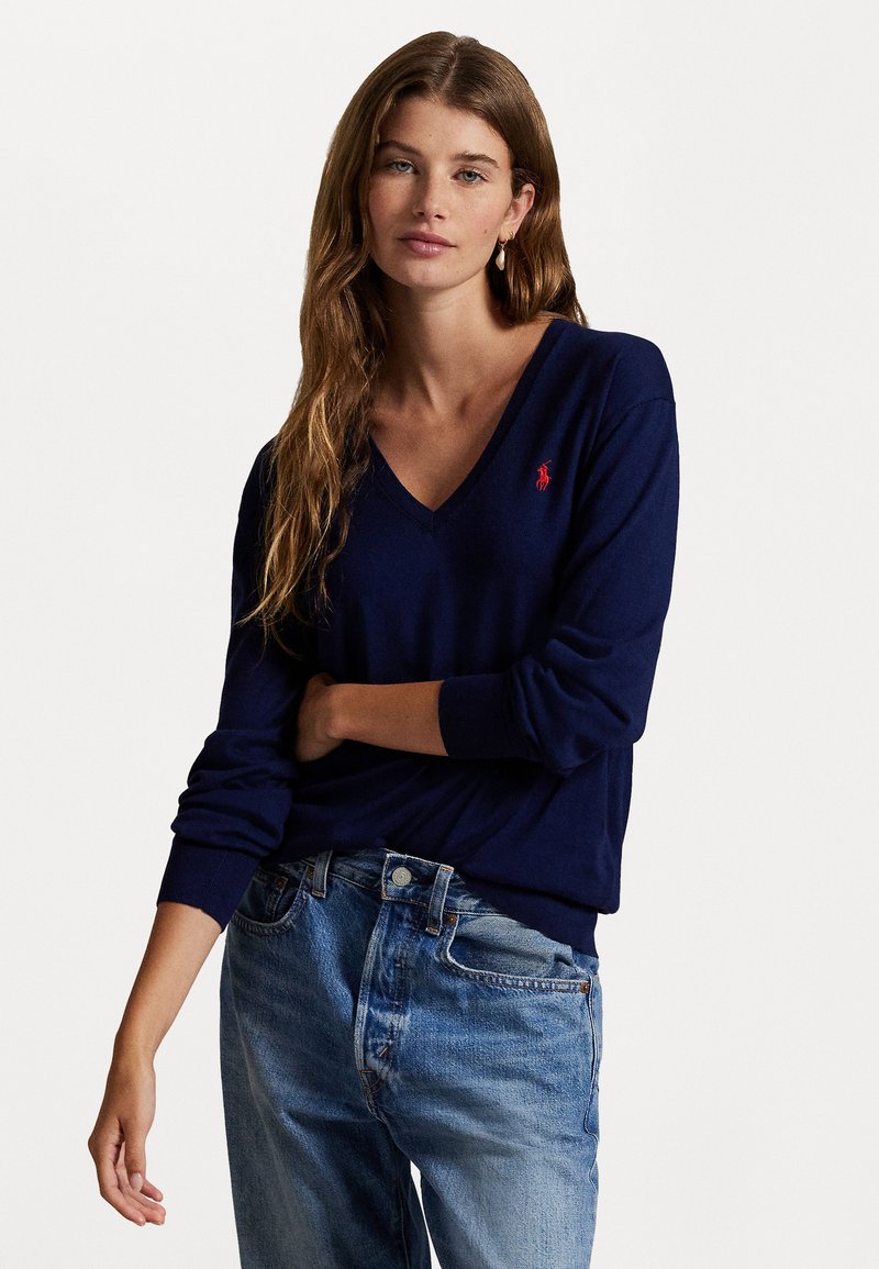 Young woman with long wavy hair wearing a navy blue V-neck sweater with a red logo and light blue denim jeans, standing against a white background.