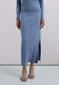 Mujer vistiendo una falda midi de punto azul con abertura lateral y sandalias de tacón alto con tiras metálicas en los tobillos, sobre un fondo liso.