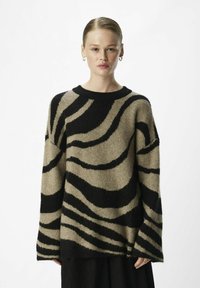 Pull beige oversize avec des rayures noires ondulées, fabriqué en matériau doux, avec un col rond et des manches longues et larges. Finition texturée.