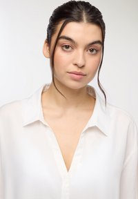 Camicia bianca con bottoni, colletto e scollatura a V; realizzata in un materiale leggero con una texture liscia e una sottile lucentezza.