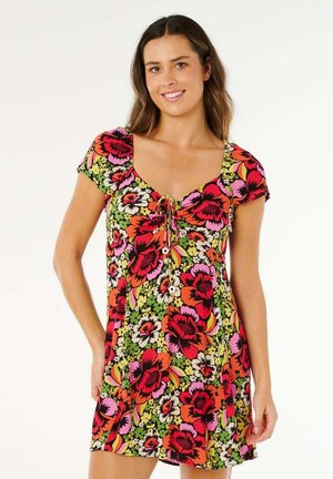 Donna con vestito a maniche corte con stampa floreale di fiori rossi, rosa, arancioni e bianchi, sorridente e in piedi di fronte a uno sfondo bianco semplice.