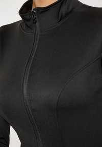Giacca sportiva nera con colletto alto e zip frontale. Il tessuto è liscio ed elastico, con cuciture sottili e un design aderente.