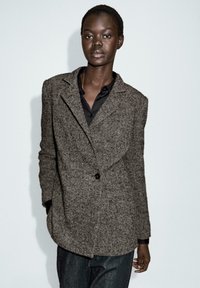 Blazer marrone con motivo a spina di pesce, con rever, chiusura a un bottone e due tasche frontali, indossato su una camicia nera e jeans blu.