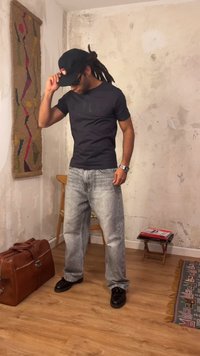Homme avec dreadlocks portant une casquette noire, des lunettes de soleil, un t-shirt noir, un jean gris et des chaussures noires se tient dans une pièce rustique avec un sac en cuir au sol.