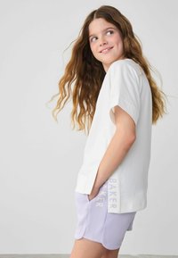 Camiseta de algodón blanca con manga corta, con un detalle en el dobladillo. Combinada con pantalones cortos lavanda que muestran la impresión "BAKER" a lo largo del costado.