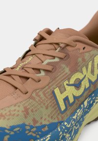 Sapato de corrida Hoka Speedgoat 6 em castanho claro com detalhes em verde e azul. Apresenta parte superior sintética texturizada, atacadores e sola com padrão.