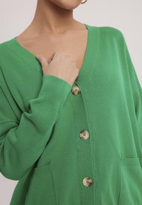 Zielony, dziergany kardigan z głębokim dekoltem w kształcie V, zapinany na guziki z przodu, posiadający dwie kieszenie i ribbowe mankiety. Miękka tekstura i luźny krój.