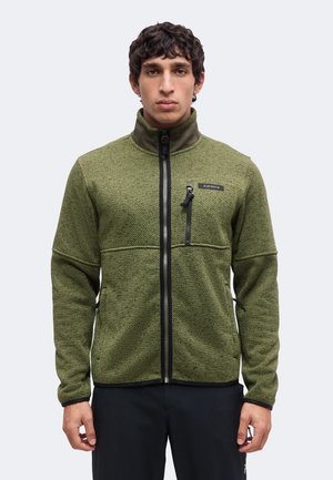 Jeune homme portant une veste zippée texturée vert olive avec un pantalon noir, debout devant un fond clair uni.
