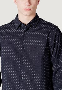 Camicia blu navy con motivi geometrici bianchi. Caratterizzata da colletto aperto, maniche lunghe e una texture del tessuto liscia e simile al cotone.