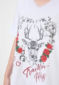 Bílý tričko s grafikou jelena obklopeného červenými růžemi a květinovými akcenty, s nápisem "Trachten-Herz" v červeném třpytu.