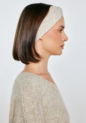 Kobieta w beżowym swetrze z dzianiny i kremowej, grubej dzianinowej opasce na głowę, z prostymi, sięgającymi ramion, brązowymi włosami, zwrócona w prawo na jednolitym tle.