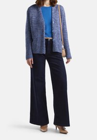 Blauer Strickcardigan über einem blauen Oberteil, kombiniert mit dunkelblauen weit geschnittenen Jeans und beigen Absätzen. Auffällige Details sind Knöpfe und ein Gürtel.