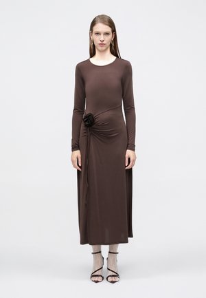 Kvinde stående iført en brun kjole med lange ærmer og et blomsterdetalje på siden, hvide blonde sokker og sorte hæle med remme mod en ensfarvet baggrund.