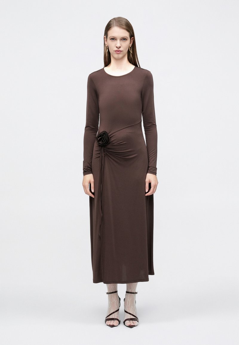 Γυναίκα που στέκεται, φορώντας μακρυμάνικο καφέ φόρεμα με λουλουδένια λεπτομέρεια στο πλάι, λευκές δαντελένιες κάλτσες και μαύρες πέδιλες με λουράκια σε απλό φόντο.