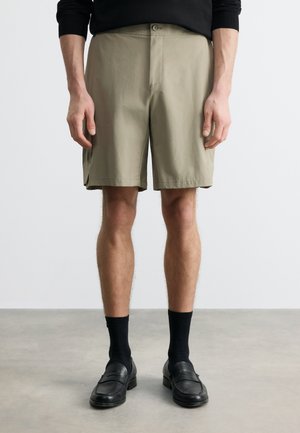 Mężczyzna w beżowych szortach za kolano, czarnych skarpetkach i czarnych mokasynach, stojący na szarym podłożu na tle jednolitej, białej ściany.