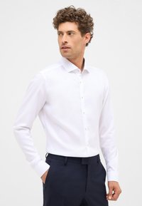 Witte lange mouwen shirt met een gestructureerde kraag, knoopsluiting aan de voorkant en subtiele textuur, gecombineerd met marineblauwe broeken. Opmerking: de knopen zijn goud.