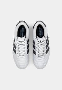 Vita sportskor med svarta ränder, texturerad läderövre, vadderad krage och vita skosnören. Har Adidas-logotypen på tungan och sidan.