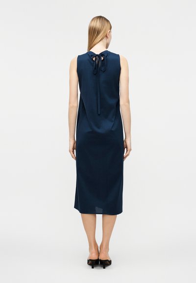 Γυναίκα με μίντι φόρεμα σε navy χρώμα χωρίς μανίκια, με φιόγκο δεμένο στο πίσω μέρος του λαιμού, στέκεται με την πλάτη προς τον θεατή, με ορατά μαύρα τακούνια.