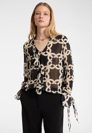Blusa nera trasparente con motivo geometrico crema, scollatura a V, dettagli arricciati lungo il davanti e polsini legati. Abbinata a pantaloni neri.