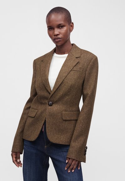 Personne aux cheveux courts portant un blazer marron à un bouton sur une chemise blanche et un jean bleu foncé, debout devant un fond uni.