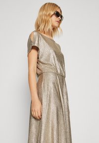 Robe dorée métallisée avec manches courtes et détail découpé aux épaules. Texture lisse, design fluide et taille cintrée.