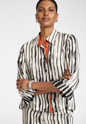 Frau mit verschränkten Armen, die eine schwarz-weiß vertikal gestreifte Bluse über einem orangefarbenen, mit Knöpfen versehenen Hemd, silberne Ohrringe und ein Armband trägt.