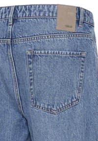 Vue arrière d'un jean en denim bleu montrant une seule poche arrière et un patch de marque en cuir beige portant l'étiquette "!SOLID" sur la ceinture.