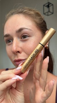 Złota tubka serum do rzęs GrandeLASH-MD z czarnym logo na opakowaniu. Gładkie metaliczne wykończenie, trzymana między palcami z długimi, różowymi paznokciami.