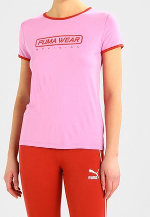 Nainen pukeutuneena vaaleanpunaiseen lyhythihaiseen Puma-paitaan, jossa on punaiset reunat, ja punaisiin Puma-housuihin yksivärisen vaalean taustan edessä.