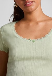 Vrouw in een lichtgroene ribgebreide top met kanten rand en een klein strikje bij de hals, gouden hoepeloorbellen zichtbaar.