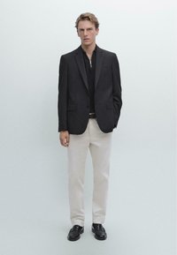 Zwarte blazer over een zwarte rits-polo, crèmekleurige broek en zwarte loafers. Gladde stof met een getailleerde fit, geen patronen of zichtbare hardware.