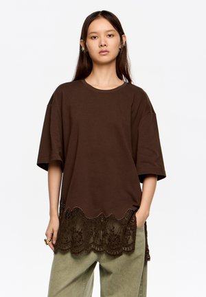 Brązowy, oversize T-shirt z falbaniastym wykończeniem z koronką na dolnym brzegu, krótkimi rękawami i okrągłym dekoltem. Materiał wydaje się miękki i przewiewny.