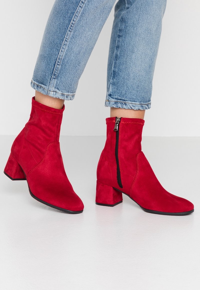 Bottes basses en daim rouge avec talons carrés et fermetures éclair latérales, portées avec un jean bleu retroussé sur un sol blanc.