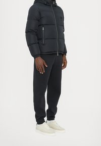 Doudoune noire avec capuche, fermeture éclair à l'avant et écusson en caoutchouc avec logo. Associée à un pantalon de survêtement foncé et des baskets de couleur claire.