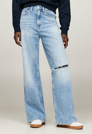 Person iført lyseblå vidde jeans med en revne på højre lår og hvide sneakers med gummisåler.