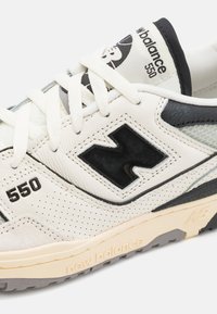 Baskets blanches et noires avec une tige en cuir perforé, présentant un grand logo "N" noir, une semelle intermédiaire crème et une semelle extérieure en caoutchouc texturé.