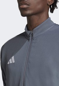 Graue Sportjacke mit hohem Kragen, Reißverschluss und dezentem adidas-Logo. Hergestellt aus strukturiertem Stoff mit glatter Oberfläche.