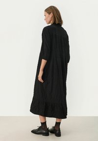 Schwarzes Maxikleid aus strukturiertem Stoff mit Dreiviertelärmeln, gestuftem Saum und lockerer Passform, kombiniert mit schwarzen Schuhen und gepunkteten Socken.