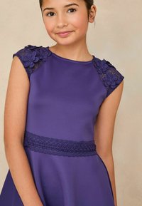 Lila Kleid mit floralen Spitzenverzierungen an den Schultern und einem strukturierten Spitzenbund. Glattes Material mit enganliegendem Oberteil und ausgestellt Rock.