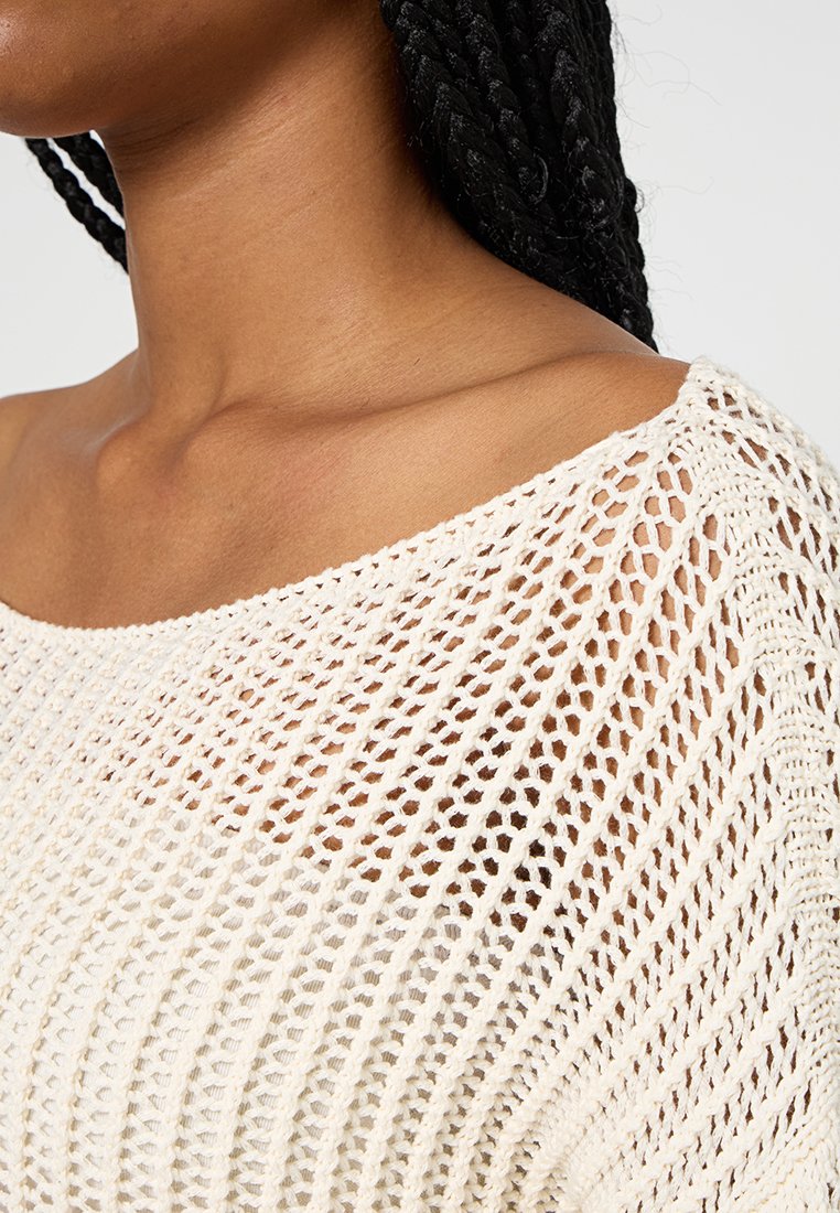 Maglione lavorato a maglia di colore crema, dal taglio morbido, con un motivo a trama aperta, scollatura ampia e tessuto testurizzato. Dettaglio della cucitura sulla spalla in evidenza.