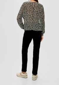 Schwarze, florale Bluse aus transparentem Stoff mit langen Ärmeln und einem Schlüsselloch-Rücken, kombiniert mit schwarzen Samthosen und beigen Sneakers mit grünen Akzenten.