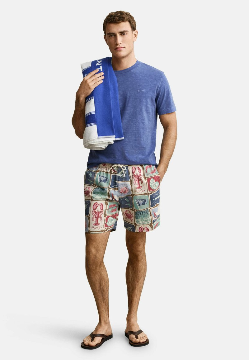 Homme portant un t-shirt bleu, un short de bain à motifs homards, tenant une serviette bleue et blanche sur l'épaule, portant des tongs noires.