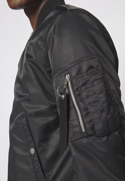 Sort bomberjakke med en glat, skinnende overflade, lynlåslomme på ærmet og en trykknaplomme foran.