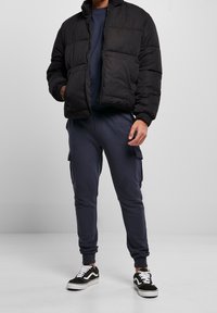 Chaqueta acolchada negra con cremallera frontal, grandes bolsillos y puños acanalados, combinada con pantalones de chándal azul marino y zapatillas Vans negras.