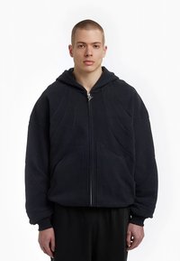Sweat à capuche noir avec fermeture éclair, fabriqué à partir d'un tissu doux. Comprend un ourlet côtelé et des poches latérales, avec des motifs de couture définis.